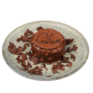 Round chocolate mousse dessert coated in milk chocolate and nut pieces, topped with a Celina chocolate disc and surrounded by chocolate shavings on a ceramic plate.