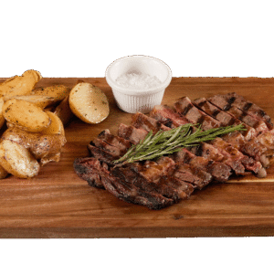 Sliced grilled ribeye steak topped with fresh rosemary, served on a wooden board with golden roasted potatoes and a ramekin of Maldon sea salt.